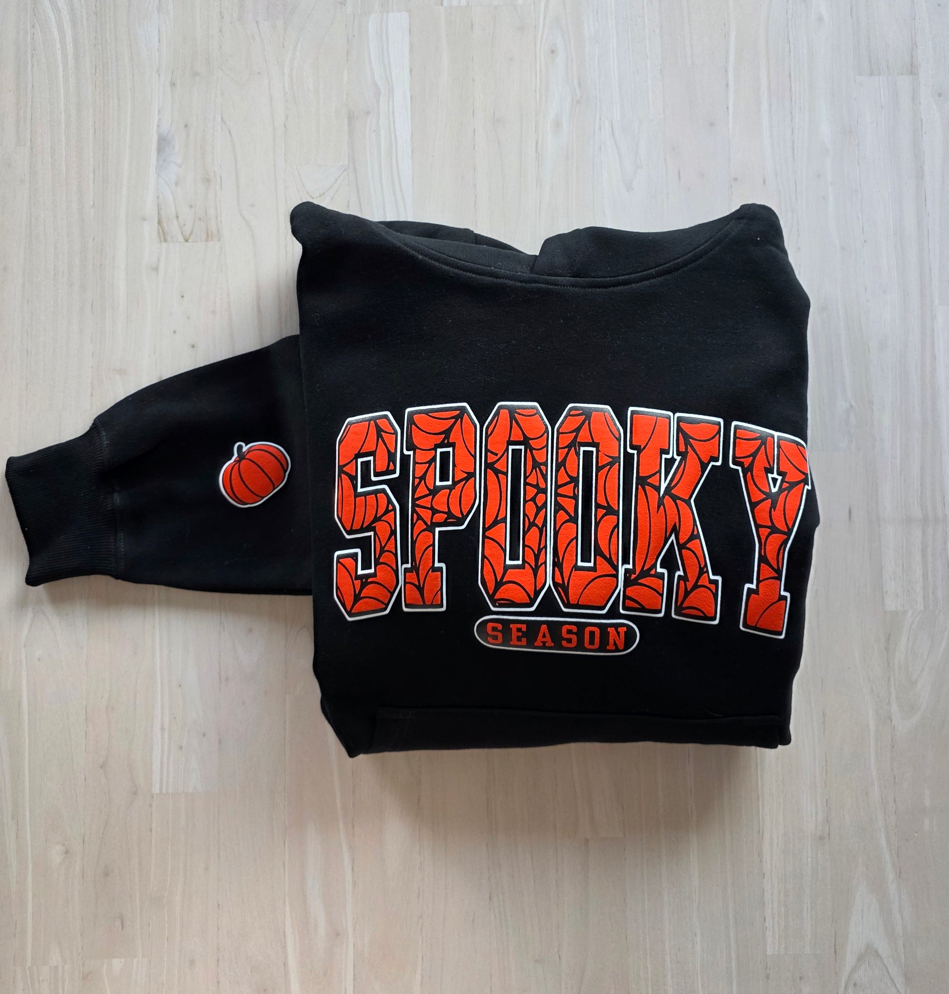 Black hoodie with 'Spooky Season' text and pumpkin graphic on a wooden surface