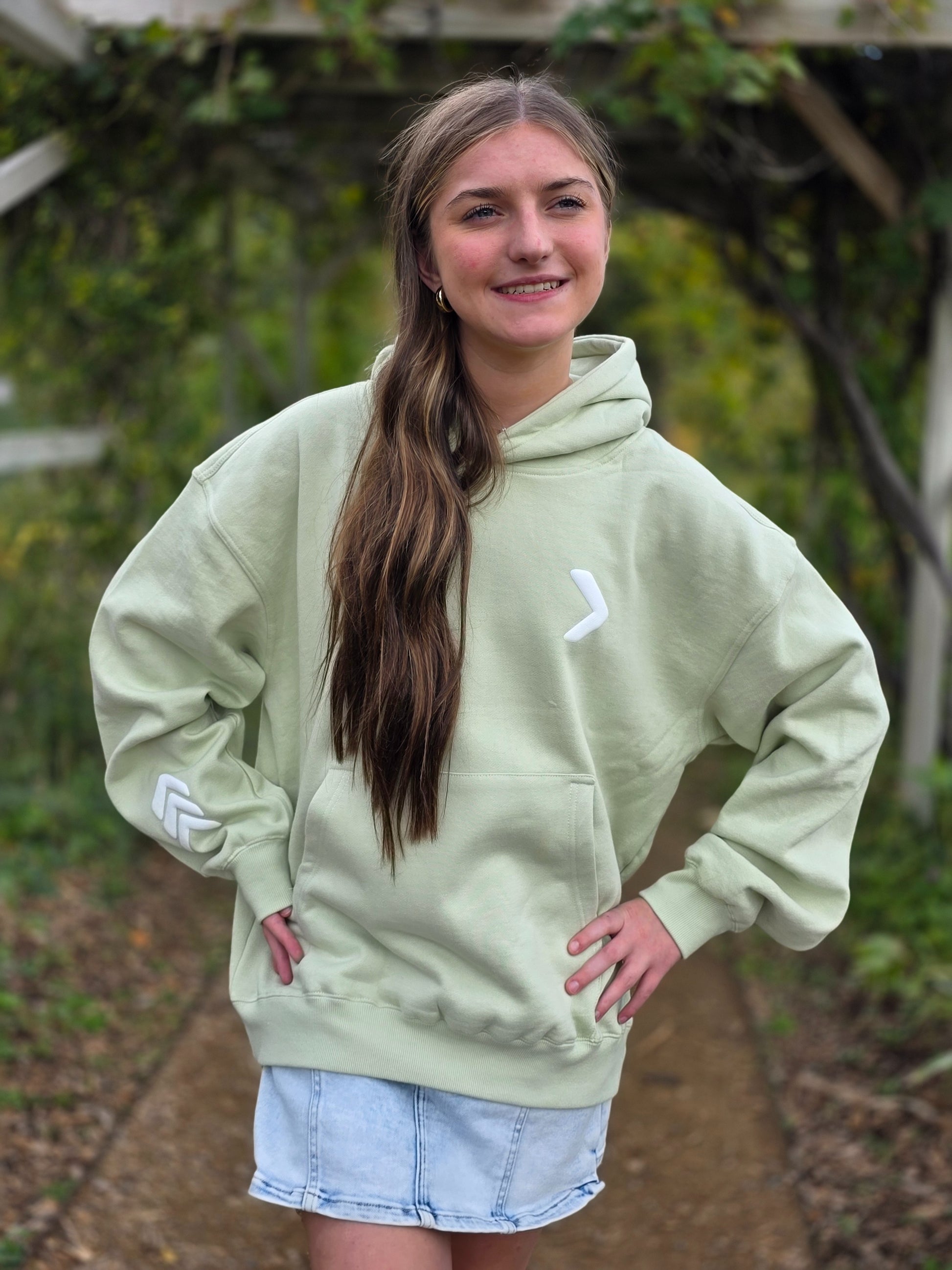 Person wearing a light green hoodie with a logo, standing outdoors with trees in the background.