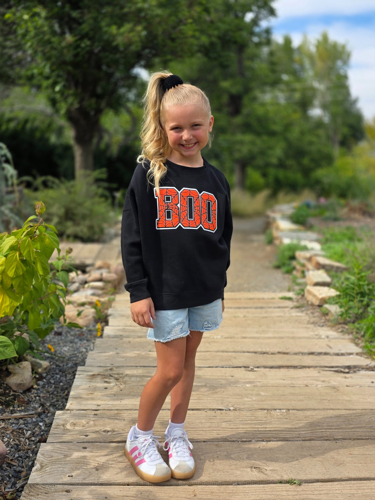 Young girl wearing a black sweatshirt with 'BOO' on it, standing on a wooden path in a garden.