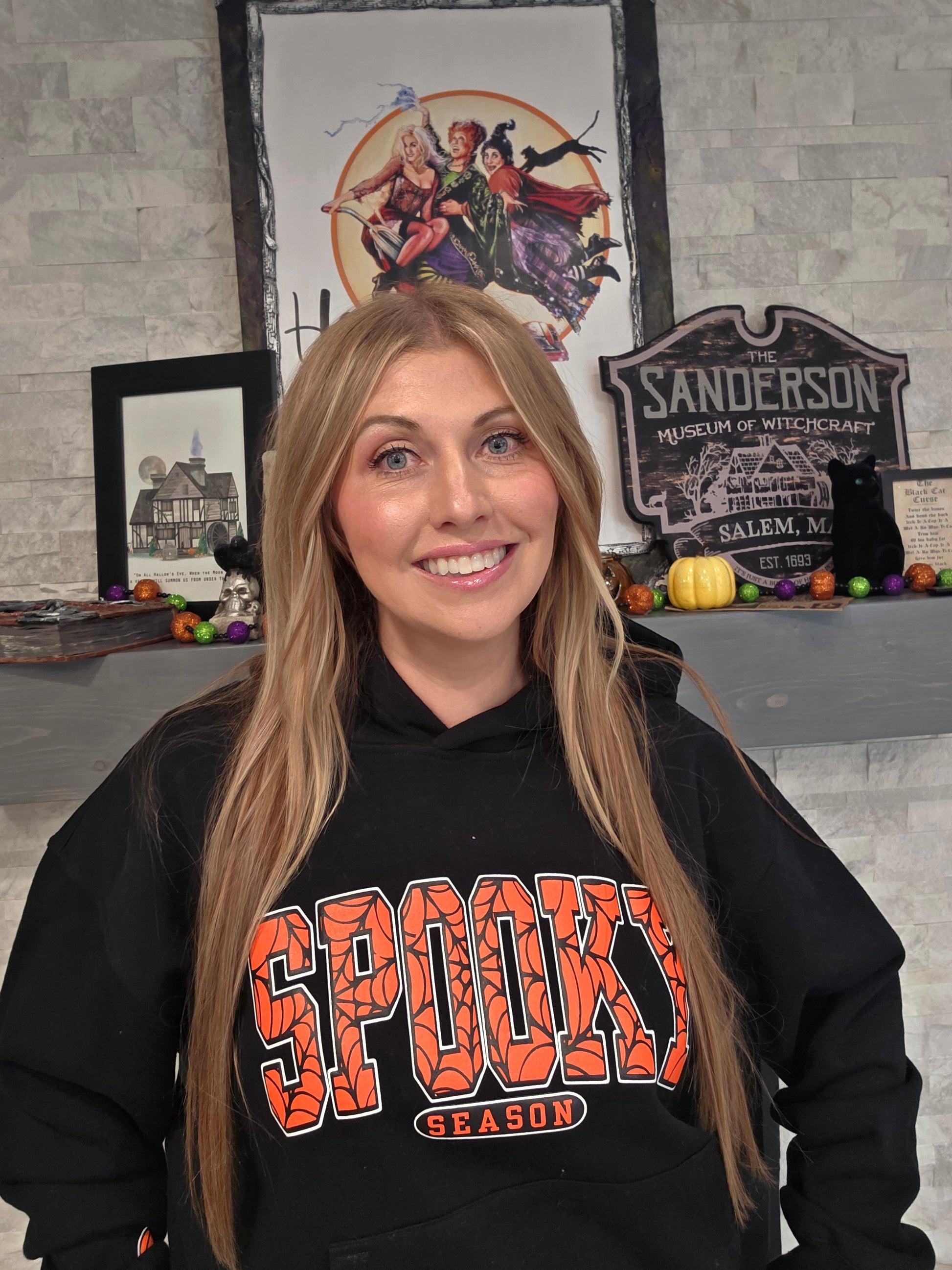Person wearing a black hoodie with 'Spooky Season' text, standing in front of Halloween-themed decorations.
