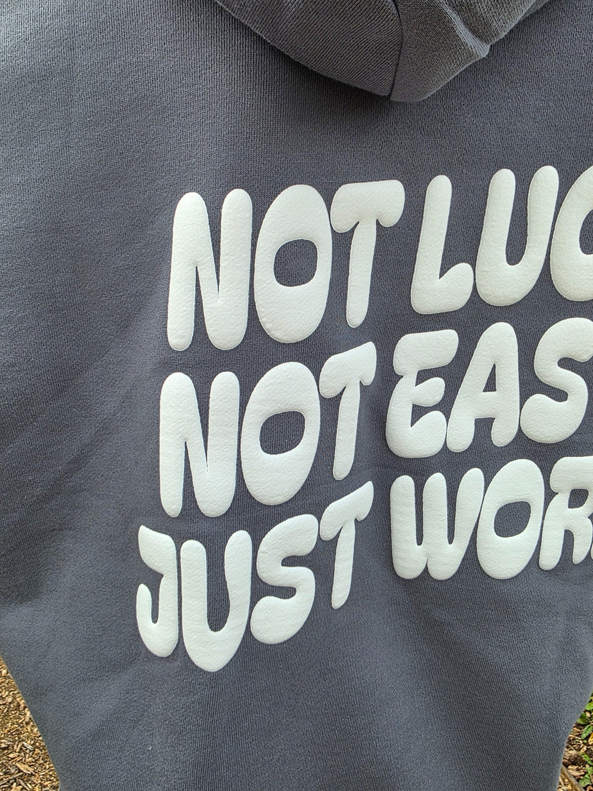 Gray hoodie with white text 'NOT LUC', 'NOT EAS', 'JUST WOR' on a blurred background
