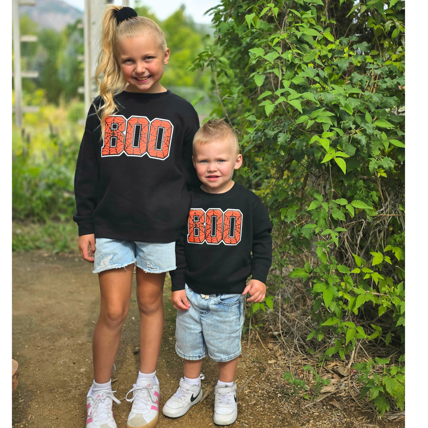 Two children wearing black sweatshirts with 'BOO' printed on them, standing outdoors.